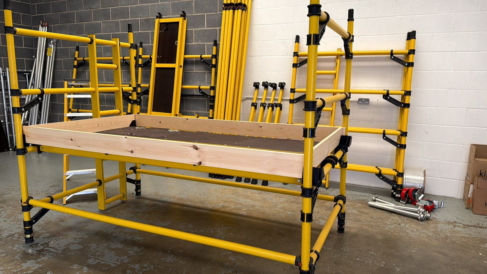 Yellow GRP fibreglass scaffold tower assembled indoors with platform, guardrails and additional GRP tower components in the background.